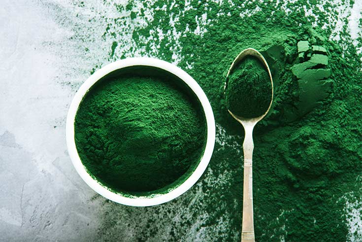 Scattered spirulina powder in bowl and spoon on concrete background.