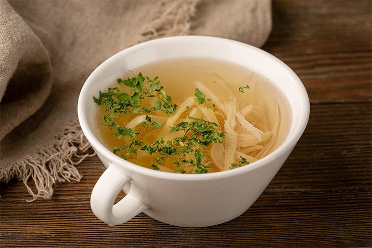 A white mug filled with vegetable bouillon and some shreds of chicken.