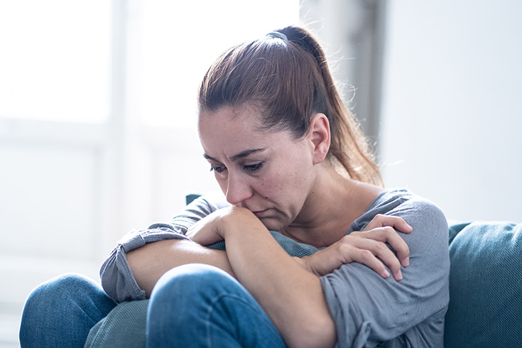 A woman sits against a bed looking sad.