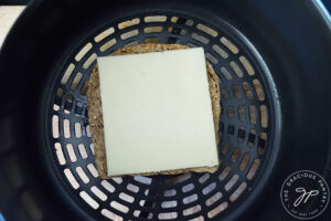 A slice of cheese laying on a slice of bread in an air fryer basket.