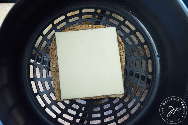 A slice of cheese laying on a slice of bread in an air fryer basket.