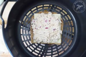 Tuna salad spread over cheese on top of a slice of bread in an air fryer basket.