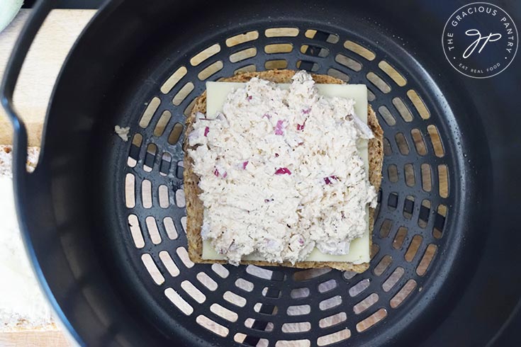 Tuna salad spread over cheese on top of a slice of bread in an air fryer basket.