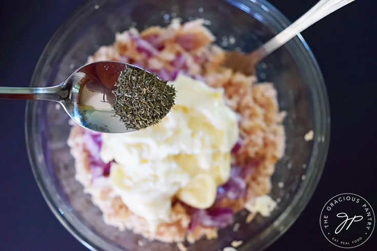 Adding dill to a glass bowl filled with tuna salad ingredients.