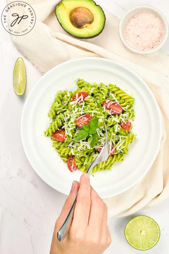 An overhead view of a female hand holding a fork reaching for a plate of Avocado Pasta.