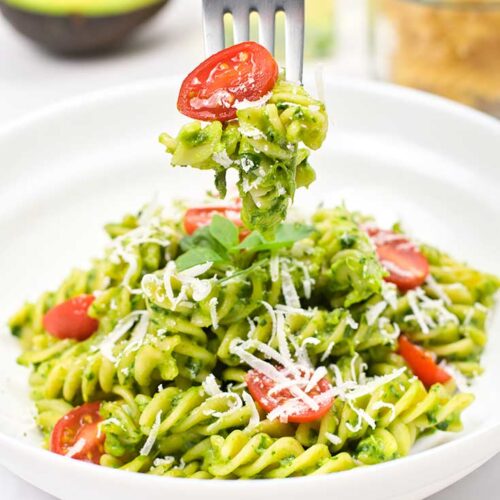 A fork holds a forkful of Avocado Pasta over a white bowl filled with it.