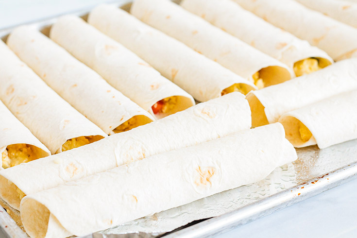Rolled Healthy Breakfast Taquitos lined up on a baking pan for baking.