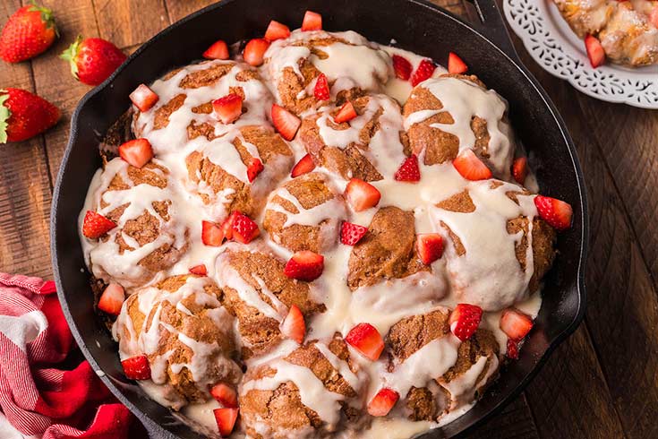 A cast iron skillet filled with french toast biscuits that have a white glaze and fresh cut strawberries garnishing the top.