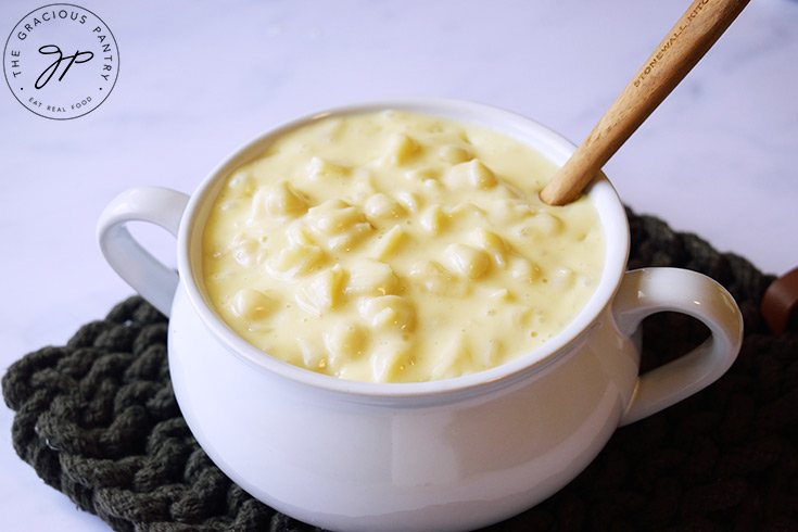 A white crock sits on a placemat filled with Homemade Mac and Cheese. A spoon rests in the pasta.