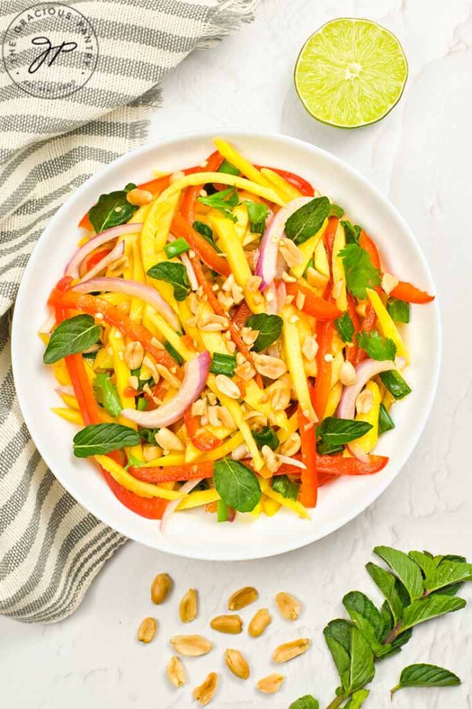 A white plate filled with Mango Salad sits on a white surface surrounded by a towel, half a lime and some peanuts.