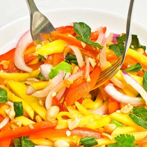 Two forks lift some mango salad out of a white bowl.