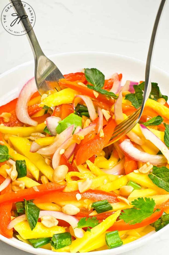 Two forks lift some mango salad out of a white bowl.