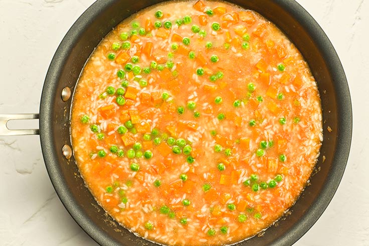 Peas and carrots added to rice and broth in a skillet.