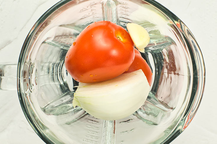 Roma tomatoes, and onion and a garlic clove sitting in a blender cup.