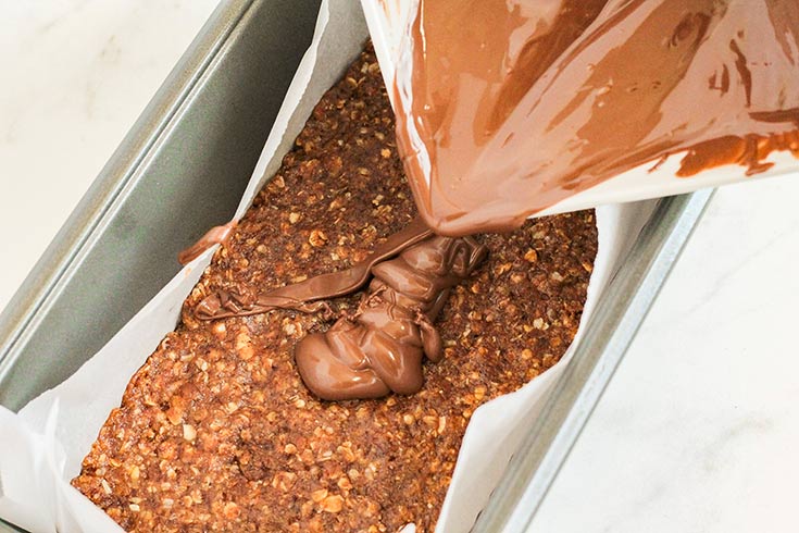 Pouring melted chocolate over the bar base in a parchment-lined loaf pan.