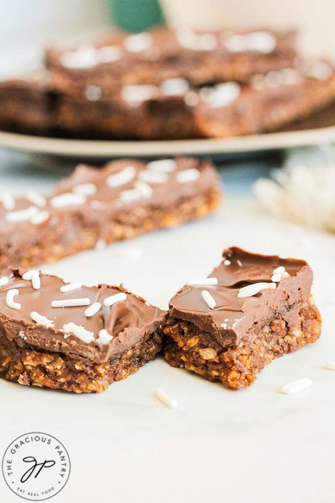 A No Bake Pumpkin Protein Bar, broken in half, laying on a white table in front of a plate of bars.