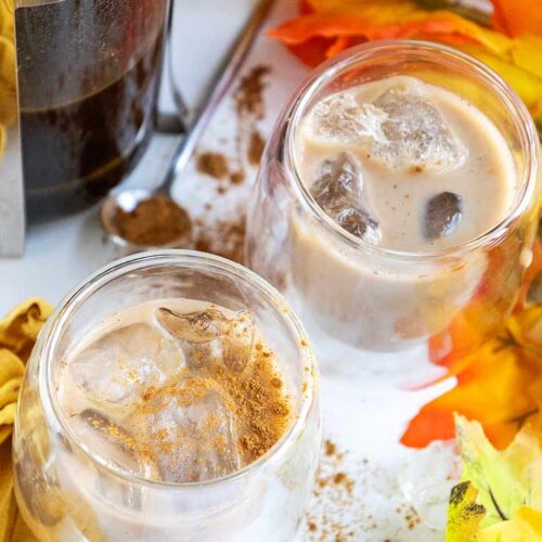 An overhead view of two cups filled with ice and Pumpkin Iced Coffee as well as a french press on a white surface with fall leaves.