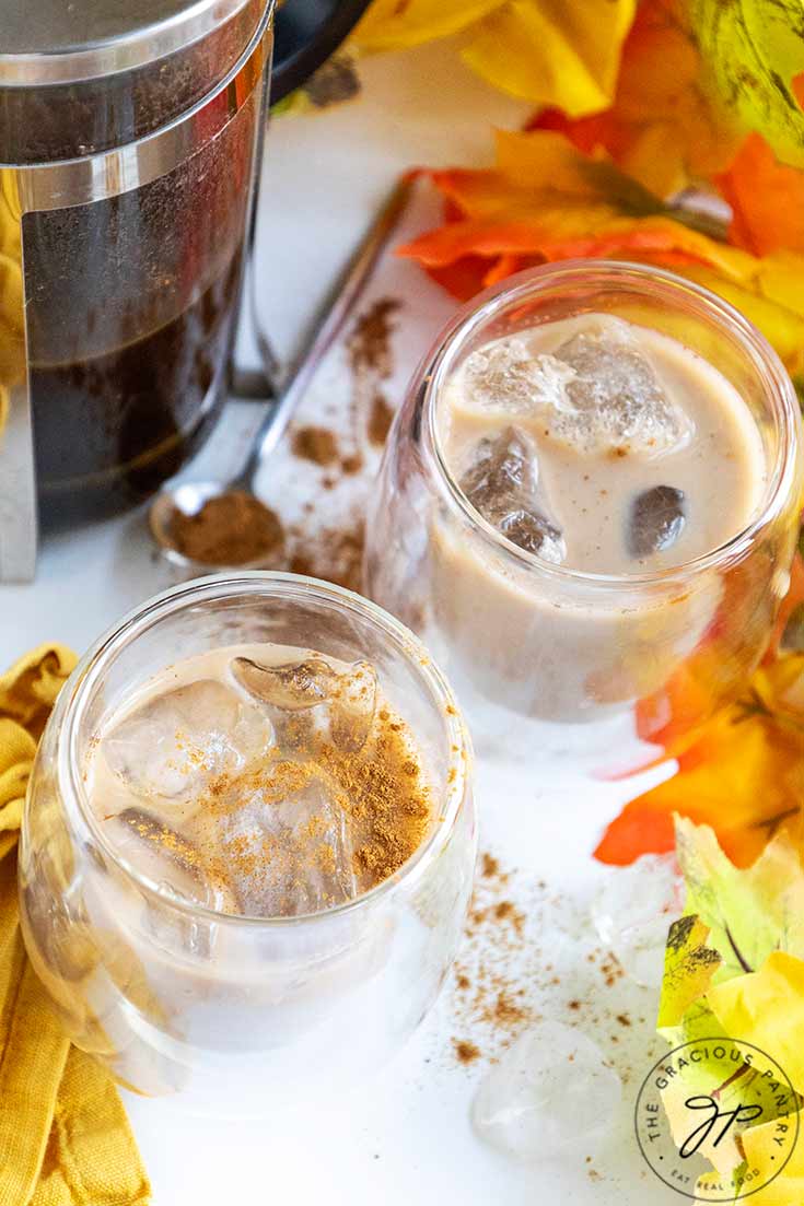 An overhead view of two cups filled with ice and Pumpkin Iced Coffee as well as a french press on a white surface with fall leaves.