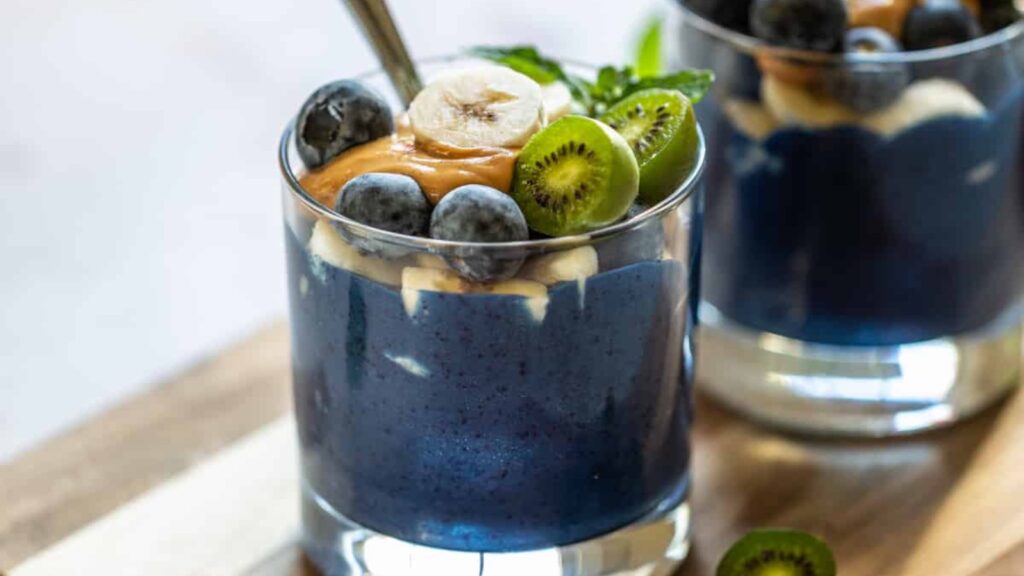 Two glasses sit filled with Spirulina Smoothie and topped with fresh fruit and a dollop of nut butter.