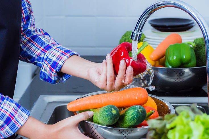 A person rinsing a red bell pepper under running water over a sink.