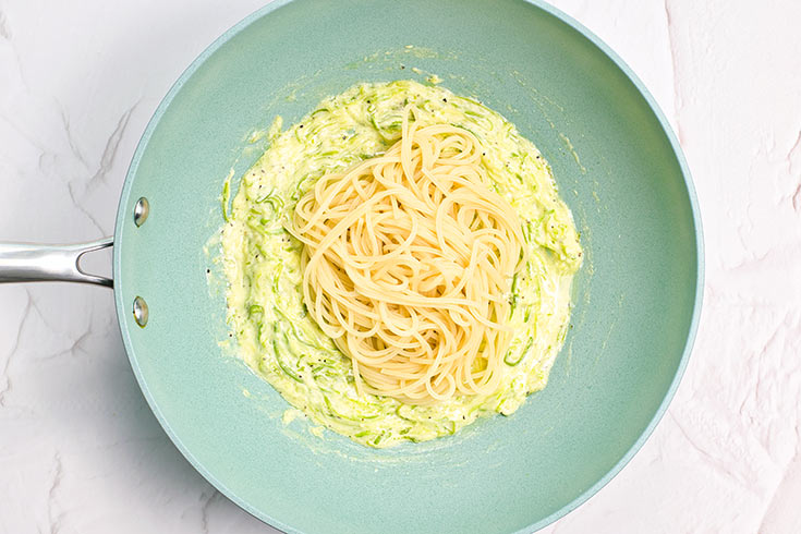 Spaghetti pasta added to creamy zucchini in a skillet.