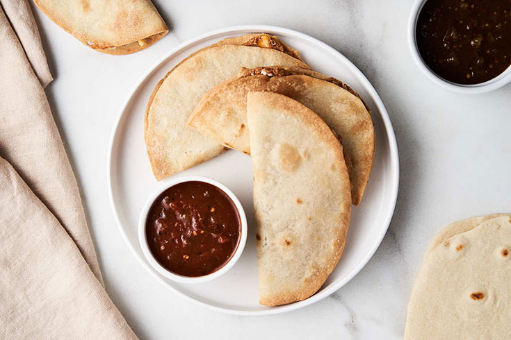 A white plate holds three air fryer quesadillas with a small bowl of sauce.