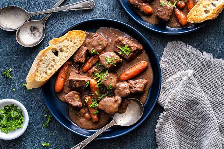 A dark blue bowl filled with beef bourguignon. A slice of bread lays on the edge of the bowl.