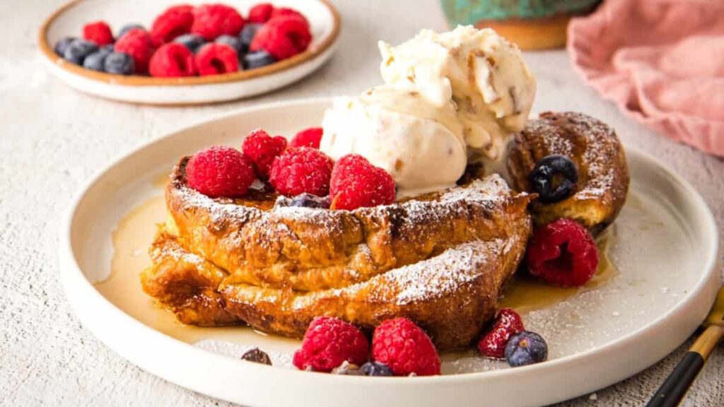 A plate holds a serving of croissant French toast bake. A plate of fresh berries sits in the background.