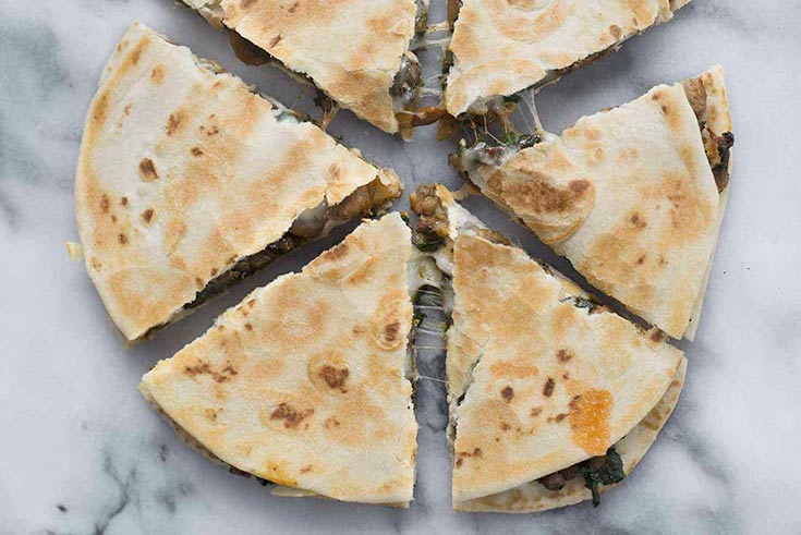 A round lentil mushroom and spinach quesadilla cut into triangles on a white marble surface.