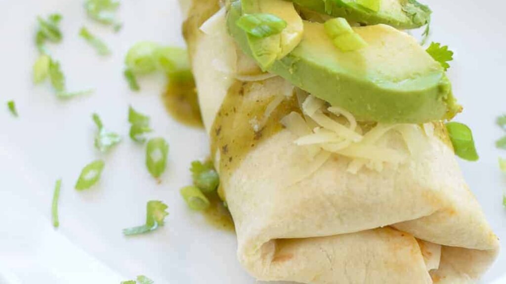 A closeup of a slow cooker breakfast burrito topped with fresh avocado, cheese and verde sauce.