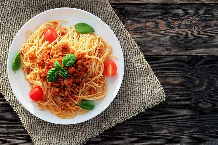 A white plate with a serving of spaghetti bolognese sitting on a wood table.