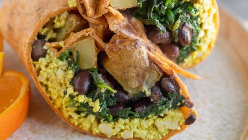 A closeup of a cut vegan breakfast burrito showing tofu, black beans, greens, and potoates.