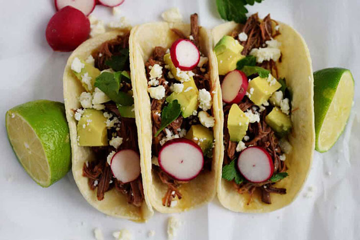 Three tacos lined up on a white surface filled with beef brisket and taco toppings.