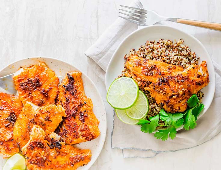 A plate with a bed of rice and a blackened cod filet sits on a white table next to a platter of blackened cod.