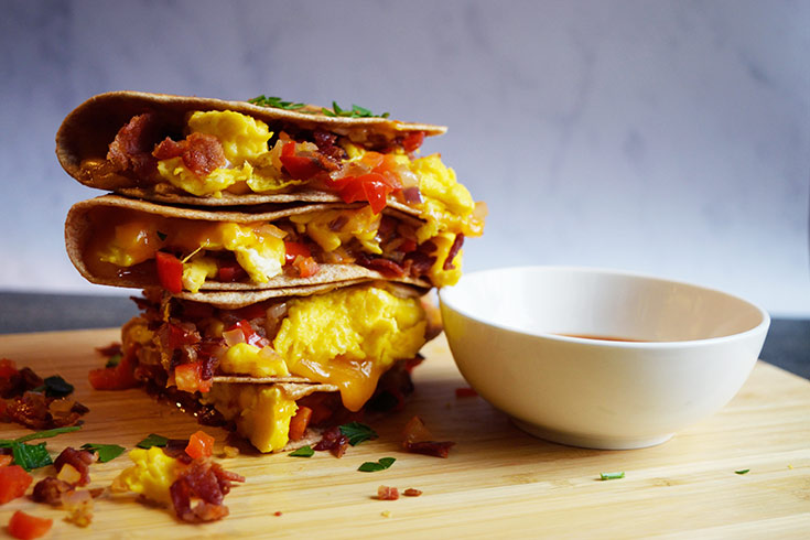 A cooked, cut and stacked Breakfast Quesadilla sitting on a cutting board next to a white bowl of salsa for dipping.