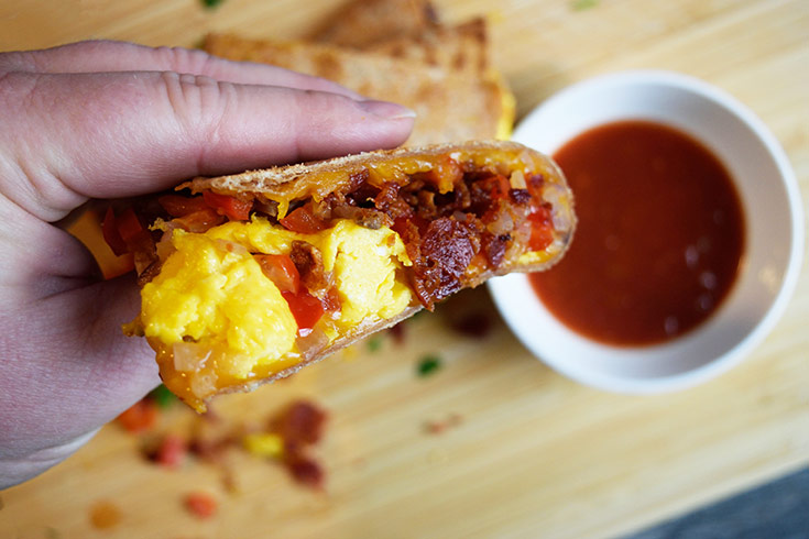A hand holds a piece of Breakfast Quesadilla up over a small, white bowl of salsa.