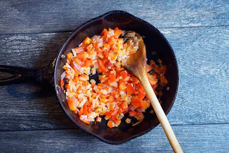 Sautéd, diced peppers and onions in a black skillet with a wooden spoon.