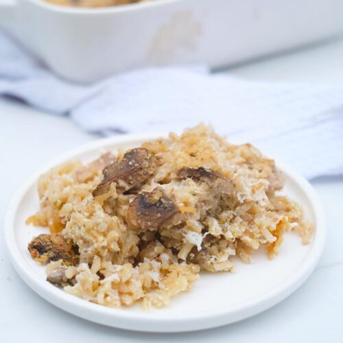 A white plate sits filled with Brown Rice Mushroom Casserole. A white casserole dish sits behind it.