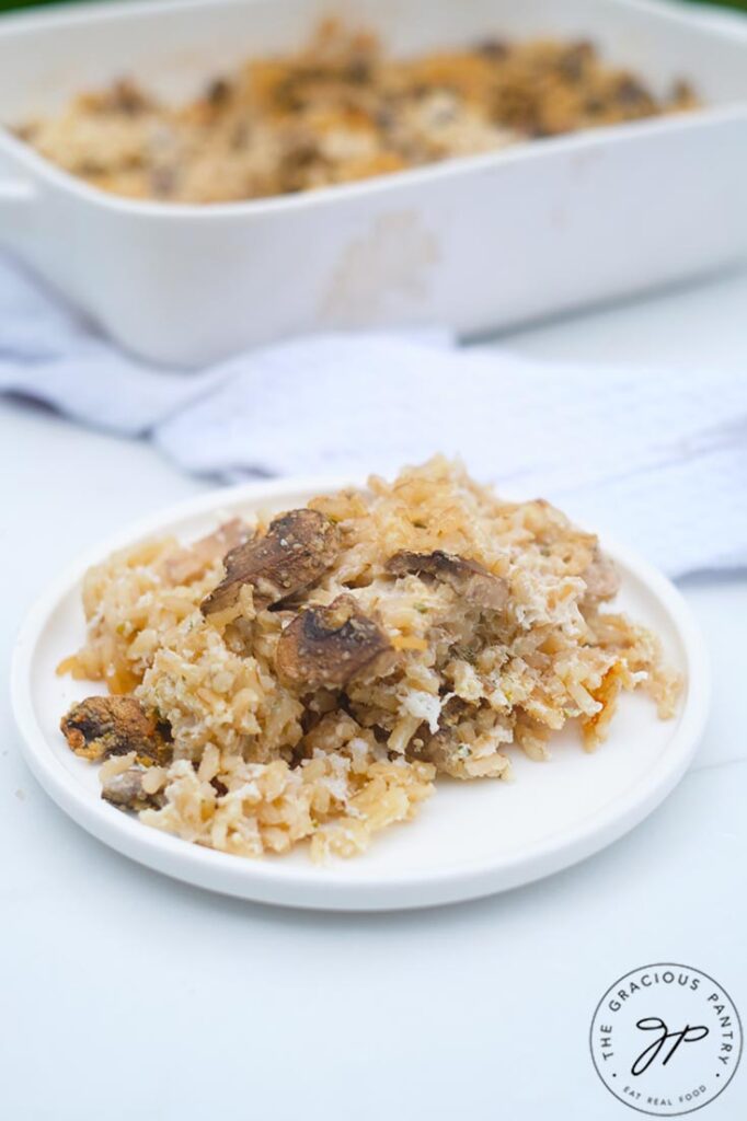 A white plate sits filled with Brown Rice Mushroom Casserole. A white casserole dish sits behind it.