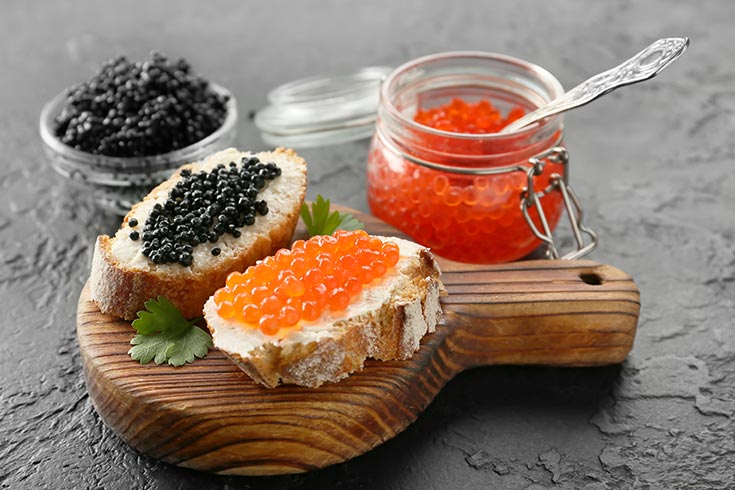 Two colors of caviar on two slices of french bread lay on a small cutting board.