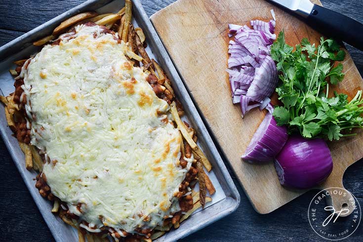 A tray of chili cheese fries sits next to a cutting board with peppers, red onion and fresh cilantro sit on it.