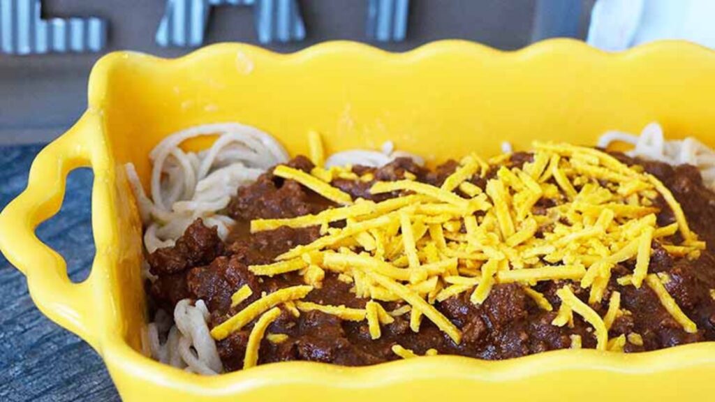 A yellow, fluted casserole dish filled with Cincinnati chili served over spaghetti pasta and topped with grated cheddar cheese.