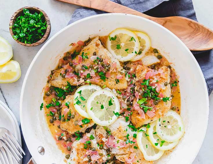 A skillet with cod piccata and lemon slices.