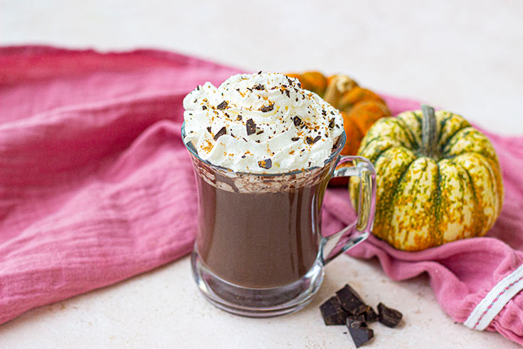 Whipped cream added to the top of a glass mug filled with pumpkin hot chocolate.