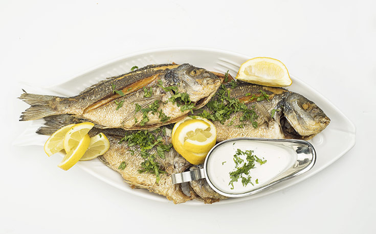 A platter of cooked, whole fish with lemon wedges and a white sauce.