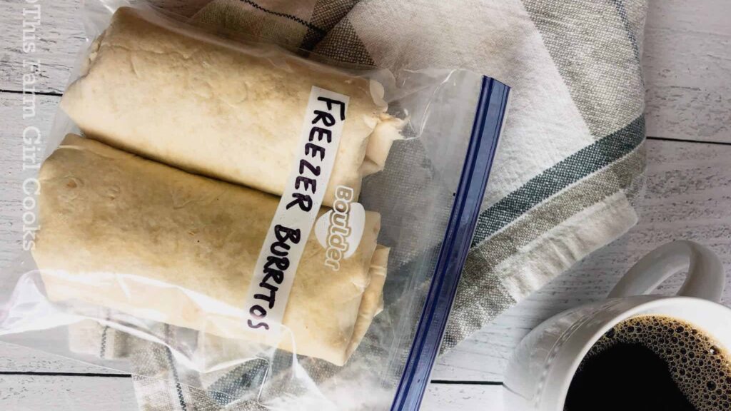 Two burritos in a freezer bag laying on a counter next to a cup of coffee.