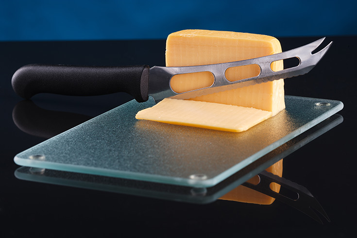 A glass cutting board with a block of cheese and a cheese knife on it.