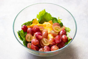 Apples and grapes added to greens in a mixing bowl.