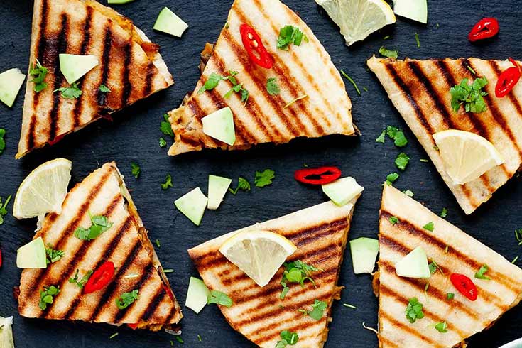 Cut hummus quesadillas laid out on a slate board with avocado pieces and lemon slices for garnish.