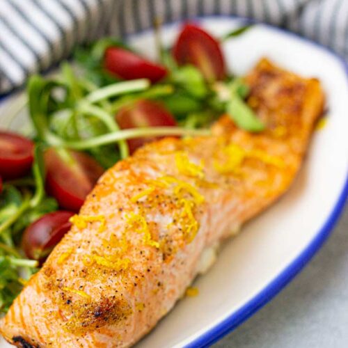 A front view of a salmon fillet on a plate with some greens and tomatoes.